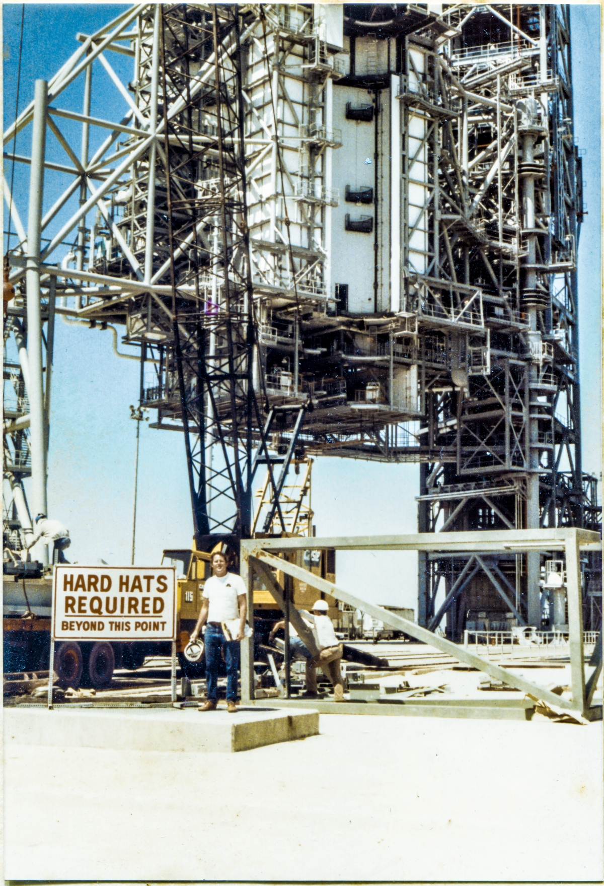 Image 055. Our earliest look at James MacLaren at Space Shuttle Launch Complex 39-B, Kennedy Space Center, Florida, after returning to the jobsite following his layoff from Sheffield Steel, and his subsequent hiring by Ivey Steel. In the far distance, the RSS and FSS loom into the sky, overflowing the frame, sensibly-complete in general appearance, but far from complete for the purposes which they were designed and constructed for, servicing the Space Shuttle, prior to launch. In the near distance, the yellow 90-ton P&H crane is being used to offload more steel from the back of a flatbed semitrailer. Union Ironworkers from Local 808 work the load from on top of the trailer and on the ground next to it, while the white-shirted truck driver lounges against the lopped-off tube steel top of the East Stair Tower. In his left hand, MacLaren grips a clipboard holding the sheets of the shipping list that came with the load of iron, with a yellow notepad behind it. In his right hand, he holds his hard hat, and if you look very close, you can also see a yellow highlighter in that hand for marking up the shipping list indicating items which have been checked and verified as being included in this delivery. On his right hip, a 25-foot tape measure that stayed with him wherever he went on the Pad Deck or on the Towers. Another day on the job. Photo by James MacLaren. Image 055. Our earliest look at James MacLaren at Space Shuttle Launch Complex 39-B, Kennedy Space Center, Florida, after returning to the jobsite following his layoff from Sheffield Steel, and his subsequent hiring by Ivey Steel. In the far distance, the RSS and FSS loom into the sky, overflowing the frame, sensibly-complete in general appearance, but far from complete for the purposes which they were designed and constructed for, servicing the Space Shuttle, prior to launch. In the near distance, the yellow 90-ton P&H crane is being used to offload more steel from the back of a flatbed semitrailer. Union Ironworkers from Local 808 work the load from on top of the trailer and on the ground next to it, while the white-shirted truck driver lounges against the lopped-off tube steel top of the East Stair Tower. In his left hand, MacLaren grips a clipboard holding the sheets of the shipping list that came with the load of iron, with a yellow notepad behind it. In his right hand, he holds his hard hat, and if you look very close, you can also see a yellow highlighter in that hand for marking up the shipping list indicating items which have been checked and verified as being included in this delivery. On his right hip, a 25-foot tape measure that stayed with him wherever he went on the Pad Deck or on the Towers. Another day on the job. Photo by James MacLaren.